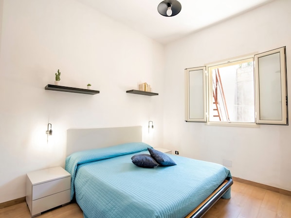 A bedroom with a bed, bedside tables, a window with a view of a construction site, and wall-mounted shelves with small plants.