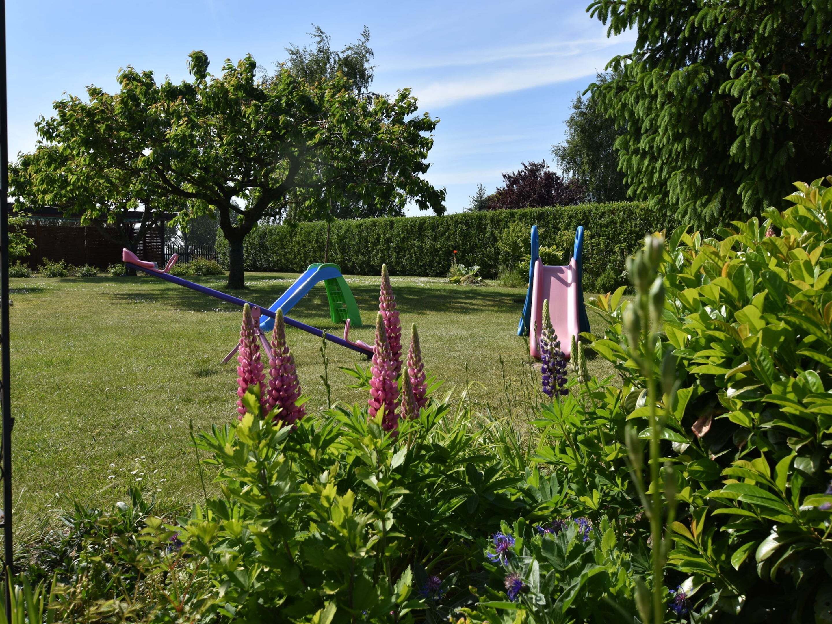 Un jardín con un parque infantil, que incluye un tobogán y columpios, rodeado de exuberante vegetación y plantas con flores.
