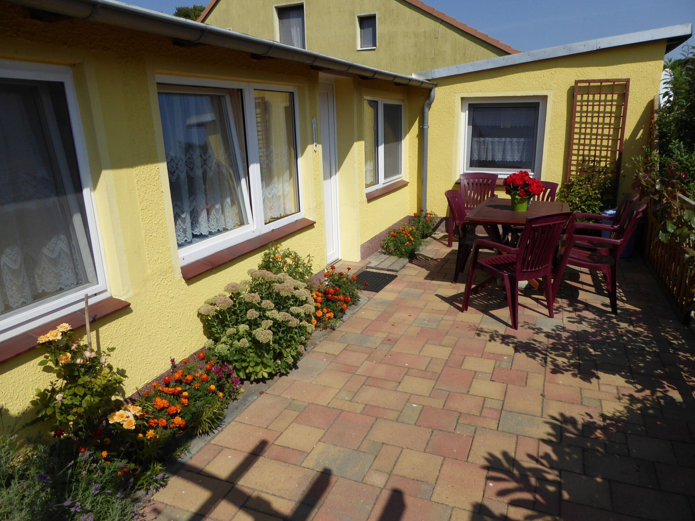 Une maison jaune avec un patio, une table et des chaises, et des parterres de fleurs.