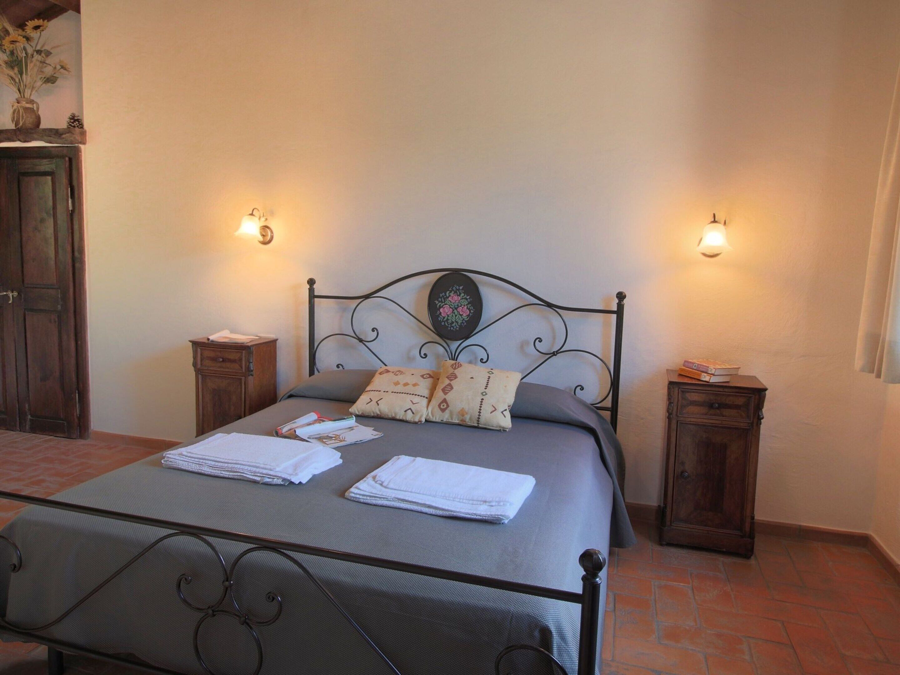 A neatly made bed with pillows and folded towels, a metal headboard, two bedside tables with lamps, and a wooden cabinet.