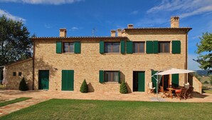 A two-story stone building with green shutters, a patio with outdoor furniture, and a clear blue sky. - Tranquil Hilltop Cottage (Montelabbate)