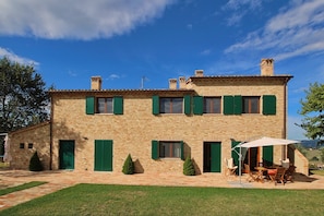 A two-story stone building with green shutters, a patio with outdoor furniture, and a clear blue sky.