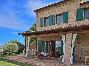 Une maison à deux étages, avec une façade en briques, des volets verts et un patio couvert aménagé avec du mobilier d’extérieur.