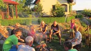 Un groupe de personnes s’est rassemblé autour d’un feu de camp dans un jardin, avec une maison en arrière-plan.
