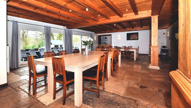 Een ruime eethoek met een grote houten tafel en stoelen, stenen vloer en een hoog houten plafond.