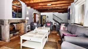 Een ruime woonkamer met een open haard, stenen vloer en een hoog houten plafond. Er zijn twee banken en een salontafel.