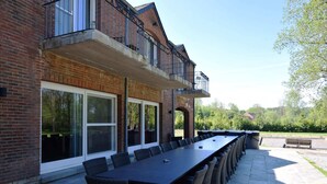 Een lange eettafel buiten, met rieten stoelen, voor een bakstenen gebouw met grote ramen en een balkon.