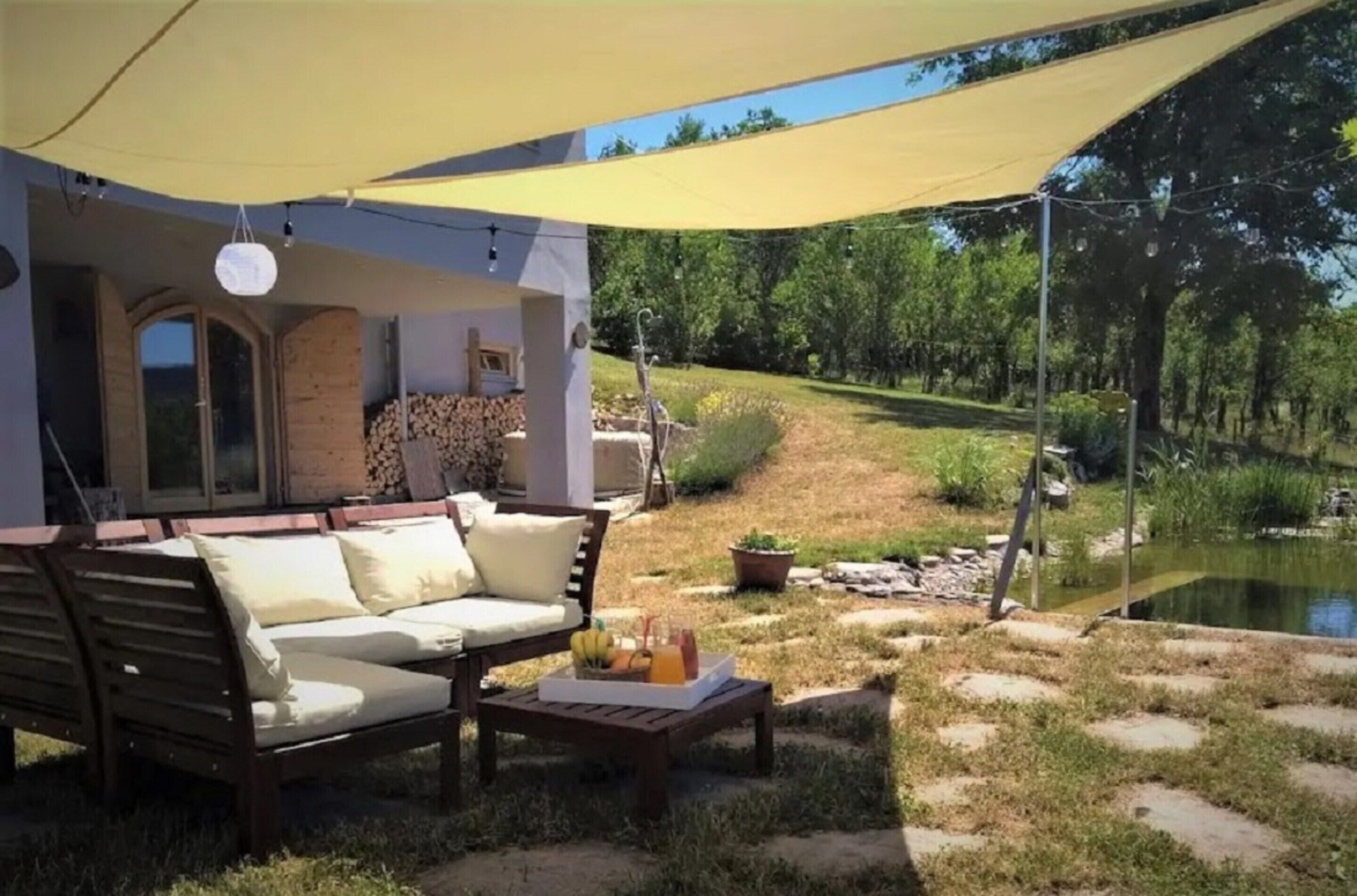 A covered outdoor seating area with a sofa and table, a small pond, and a garden with trees and a path.