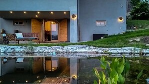 A modern building with a glass door entrance, a wooden deck, and a pond reflecting the structure. - Comfy Holiday Home in Bunjevci With Hot Tub (Vrbovsko)