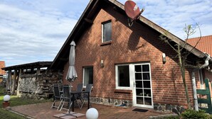 A brick house with a sloped roof, a satellite dish, a covered patio with a table and chairs, and a small garden area.