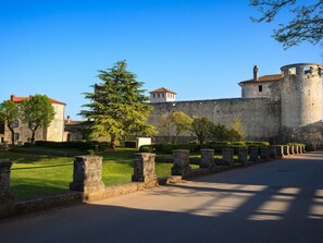 Un mur de pierre ancien, surmonté d’une tour ronde, une zone herbeuse avec un grand arbre et une route pavée.