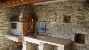 A stone-built outdoor kitchen area with a built-in oven, a hanging rack with tools, and a stone bench. - Stone Holiday Home in Peresiji With Pool (Svetvincenat)