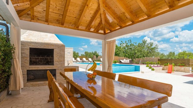 An outdoor patio with a wooden table and benches, a stone fireplace, and a pool area with lounge chairs. - Modern Home in Quiet Pristeg (Benkovac)