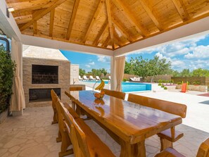 An outdoor patio with a wooden table and benches, a stone fireplace, and a pool area with lounge chairs. - Modern Home in Quiet Pristeg (Benkovac)