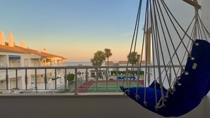 Un balcon avec un hamac suspendu, une vue sur un court de tennis et des bâtiments de style architectural méditerranéen.