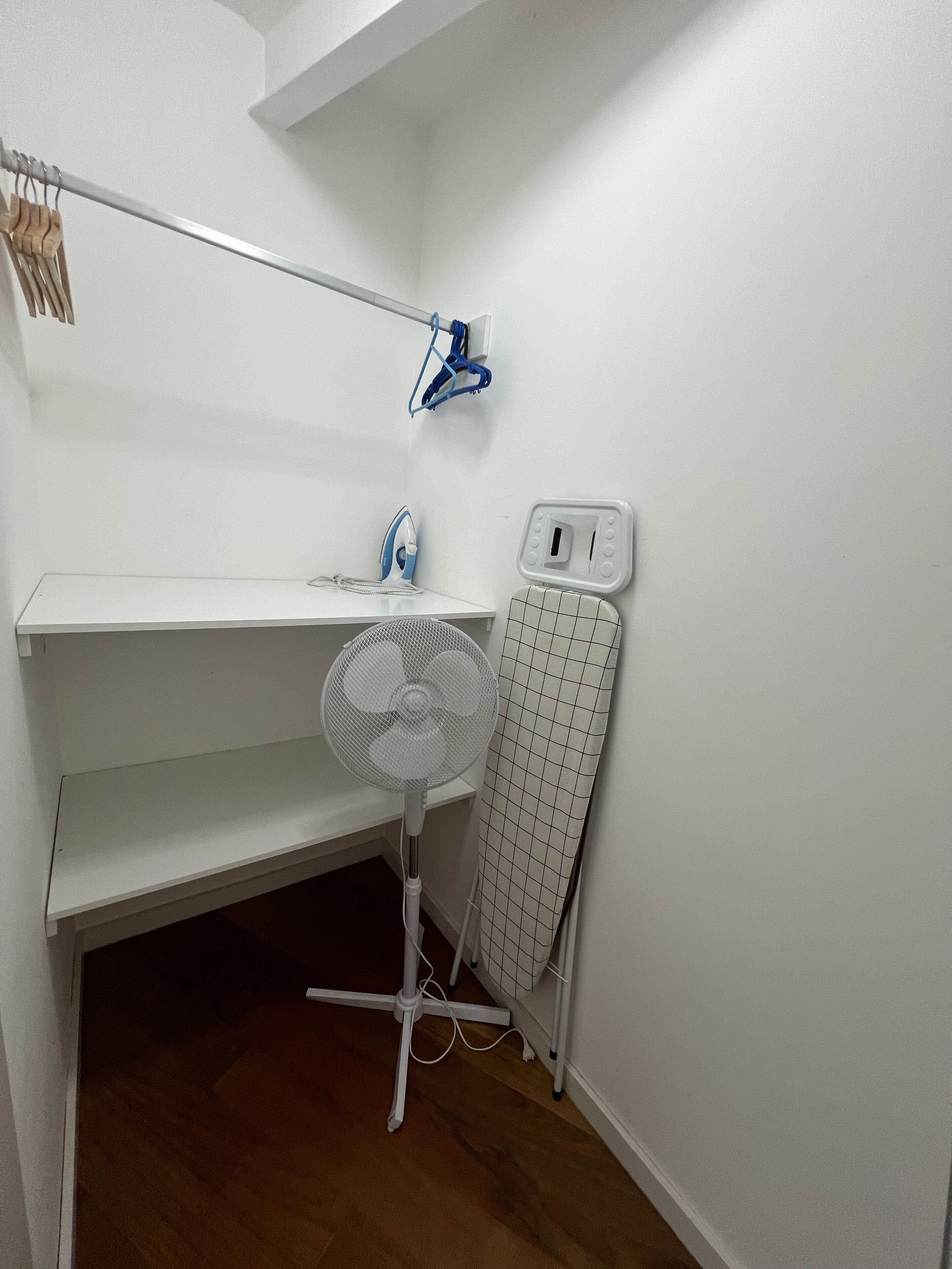 Un pequeño cuarto de servicio blanco con estante, tabla de planchar, ventilador y un enchufe en la pared.