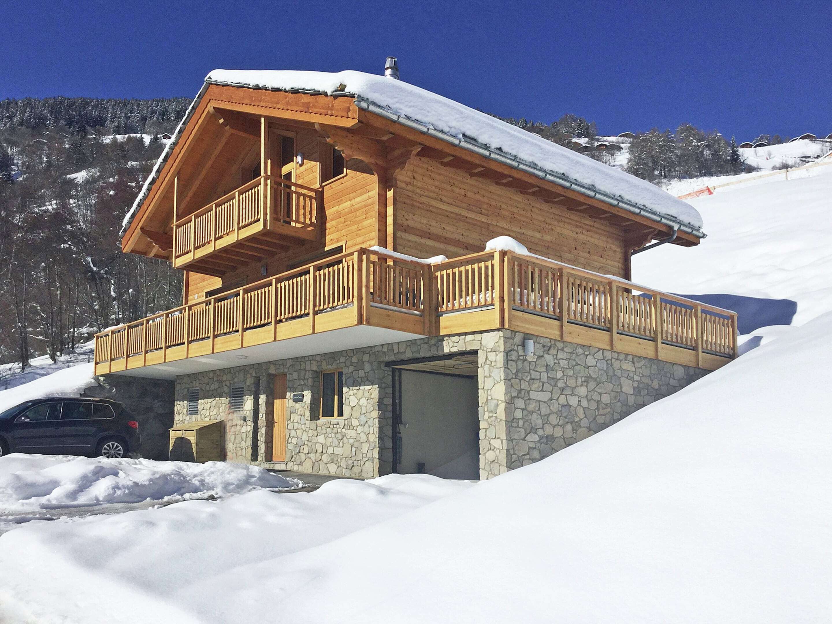 A wooden chalet with a balcony, stone foundation, and a car parked in front.