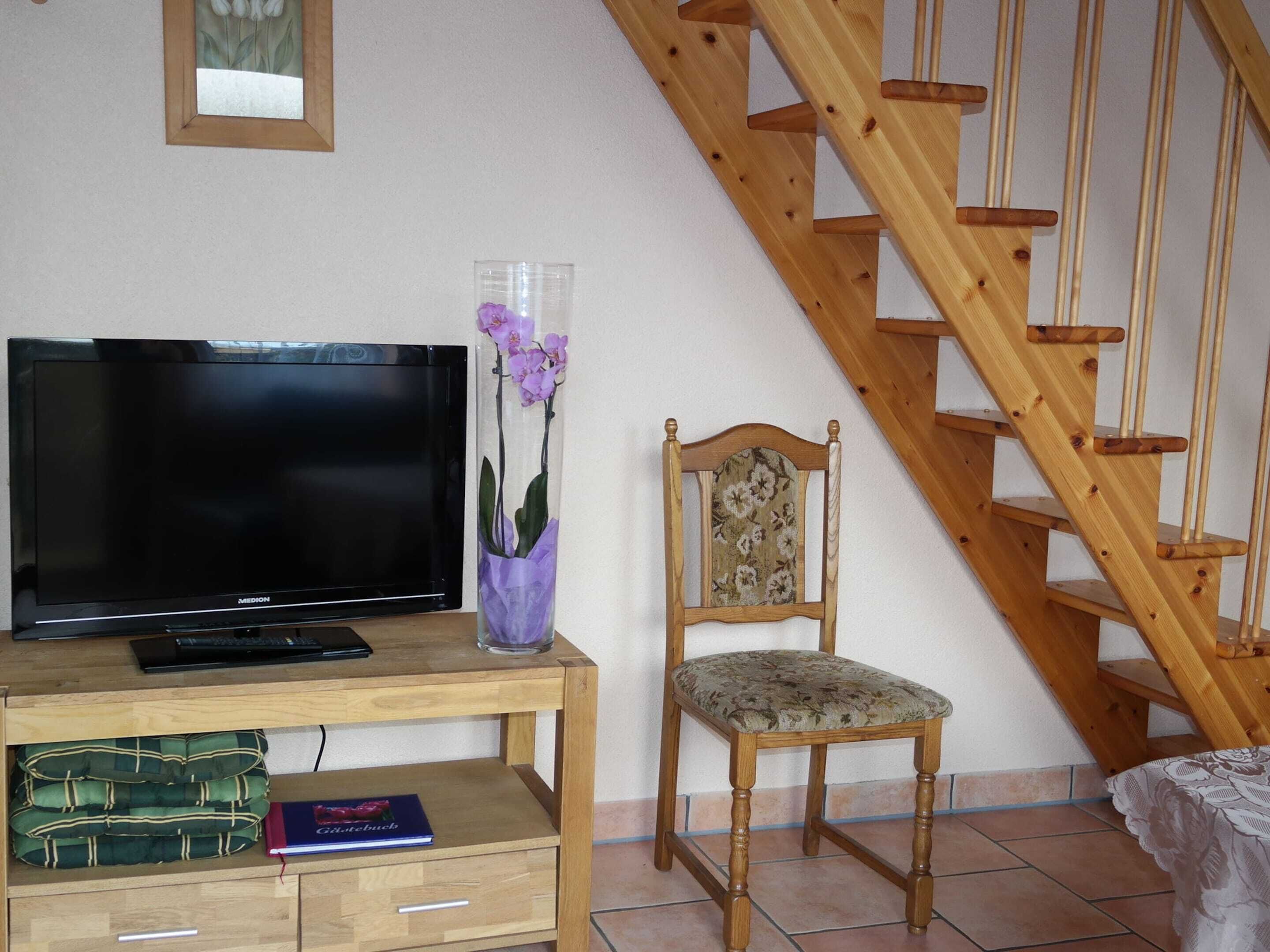 A living room with a wooden TV stand, a flat-screen TV, a chair, a vase with flowers, and a wooden staircase.