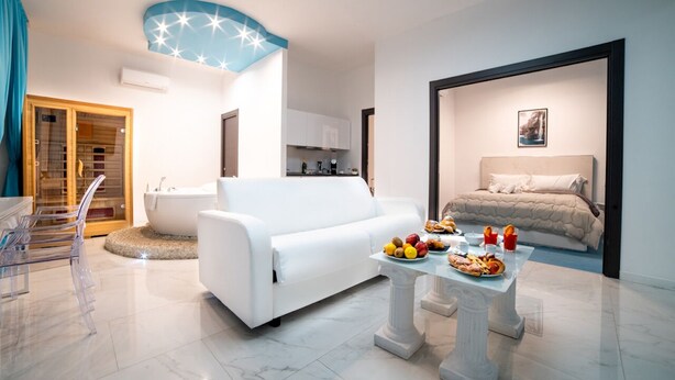 Una sala de estar moderna con un sofá blanco, una mesa de centro de vidrio con frutas, una bañera y una cama con cabecera.