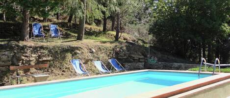A rectangular swimming pool with a stone border, surrounded by a stone wall and trees. There are two lounge chairs with blue cushions and a wooden bench.