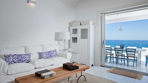 Ein modernes Wohnzimmer mit einem weißen Sofa, gemusterten Kissen, einem Holztisch und Blick aufs Meer durch eine Glastür.