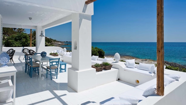 Eine weiße Terrassengruppe im Freien mit Tisch, Stühlen und einer Sitzecke mit Kissen, mit Blick auf das Meer.