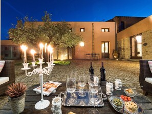 Un área de comedor al aire libre con una mesa dispuesta para dos personas, incluyendo copas de vino, una bandeja con aceitunas y pan, y un candelabro.