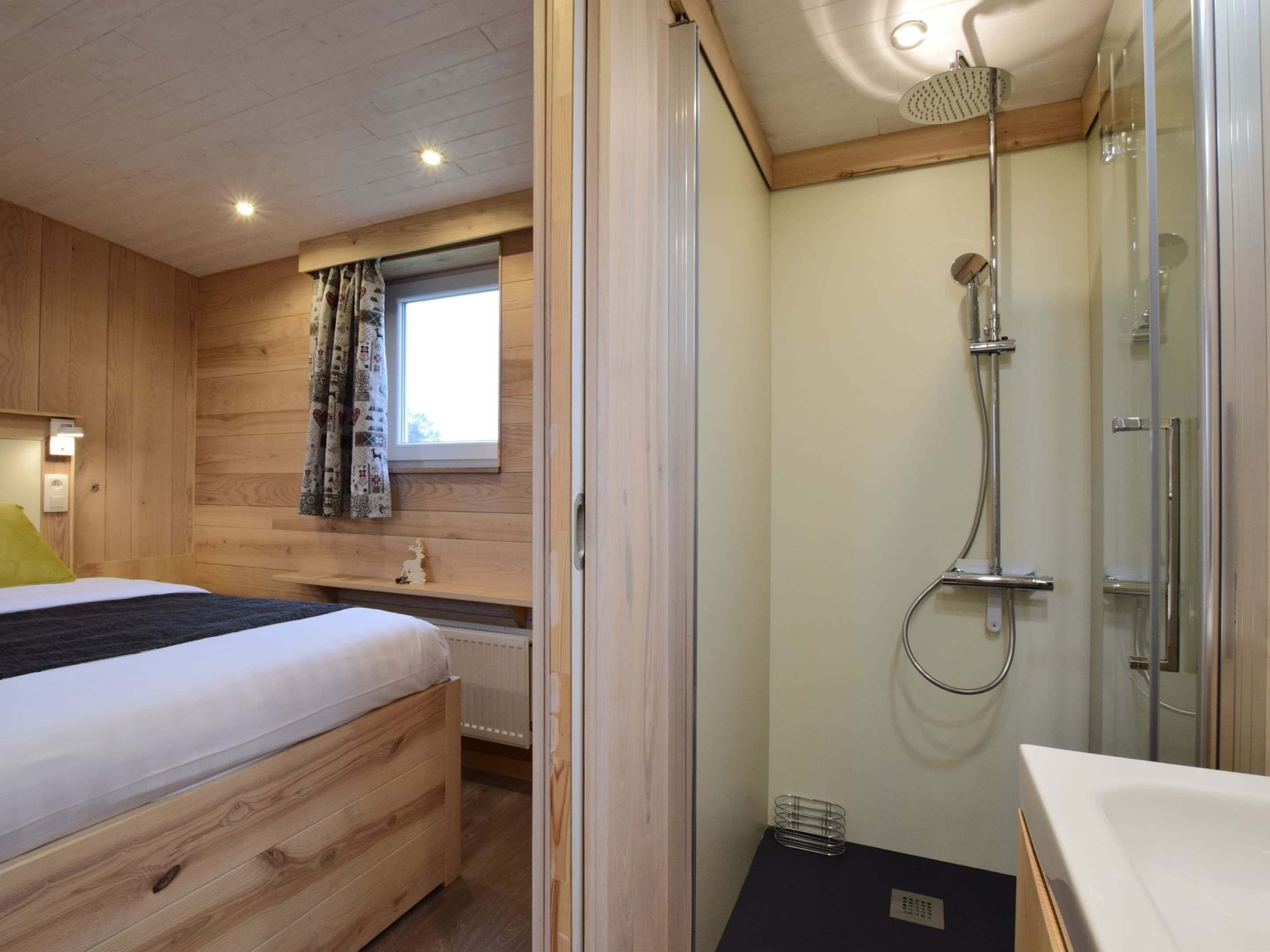Una habitación de madera con una cama, una ventana con cortinas, una ducha con mampara de cristal y un radiador.