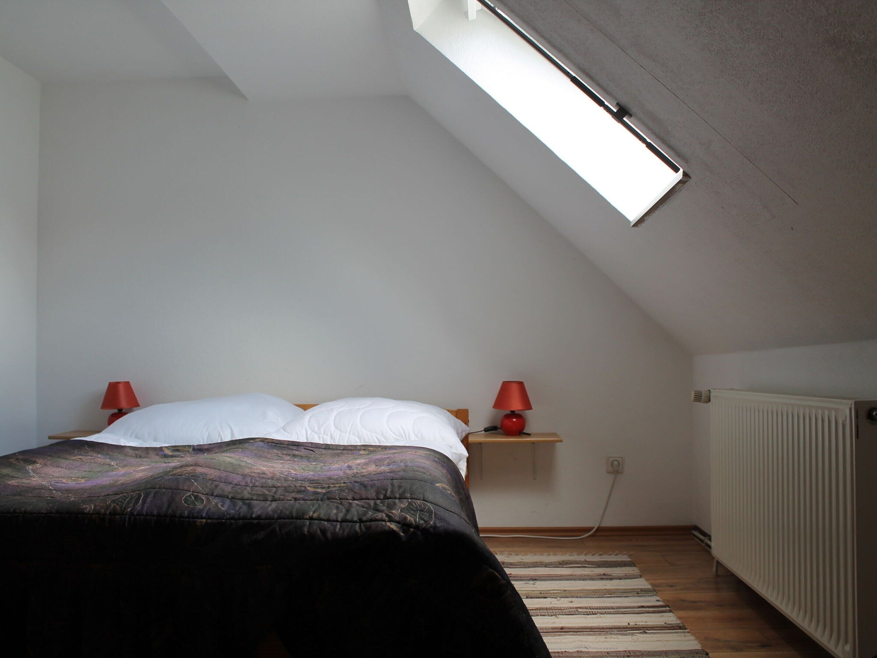 A bedroom with a sloped ceiling, a bed with white and black bedding, two red bedside lamps, a small wooden nightstand, and a radiator.
