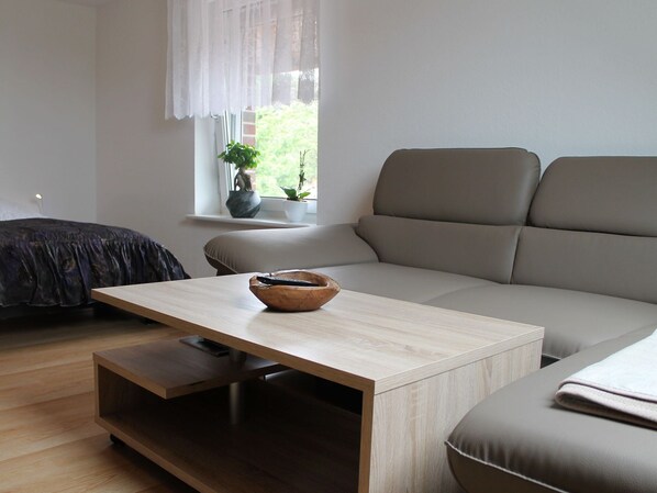 Uma sala de estar moderna com uma mesa de centro de madeira clara, um sofá cinza e uma cama de cor escura.