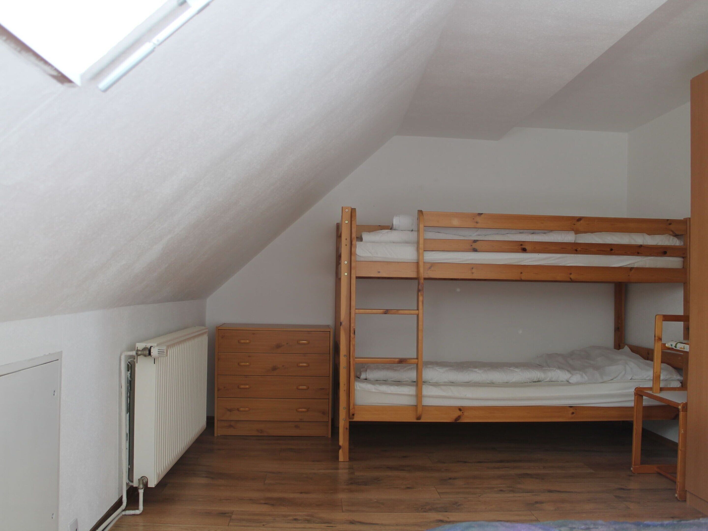 A dormitory room with a bunk bed, a wooden dresser, and a radiator.
