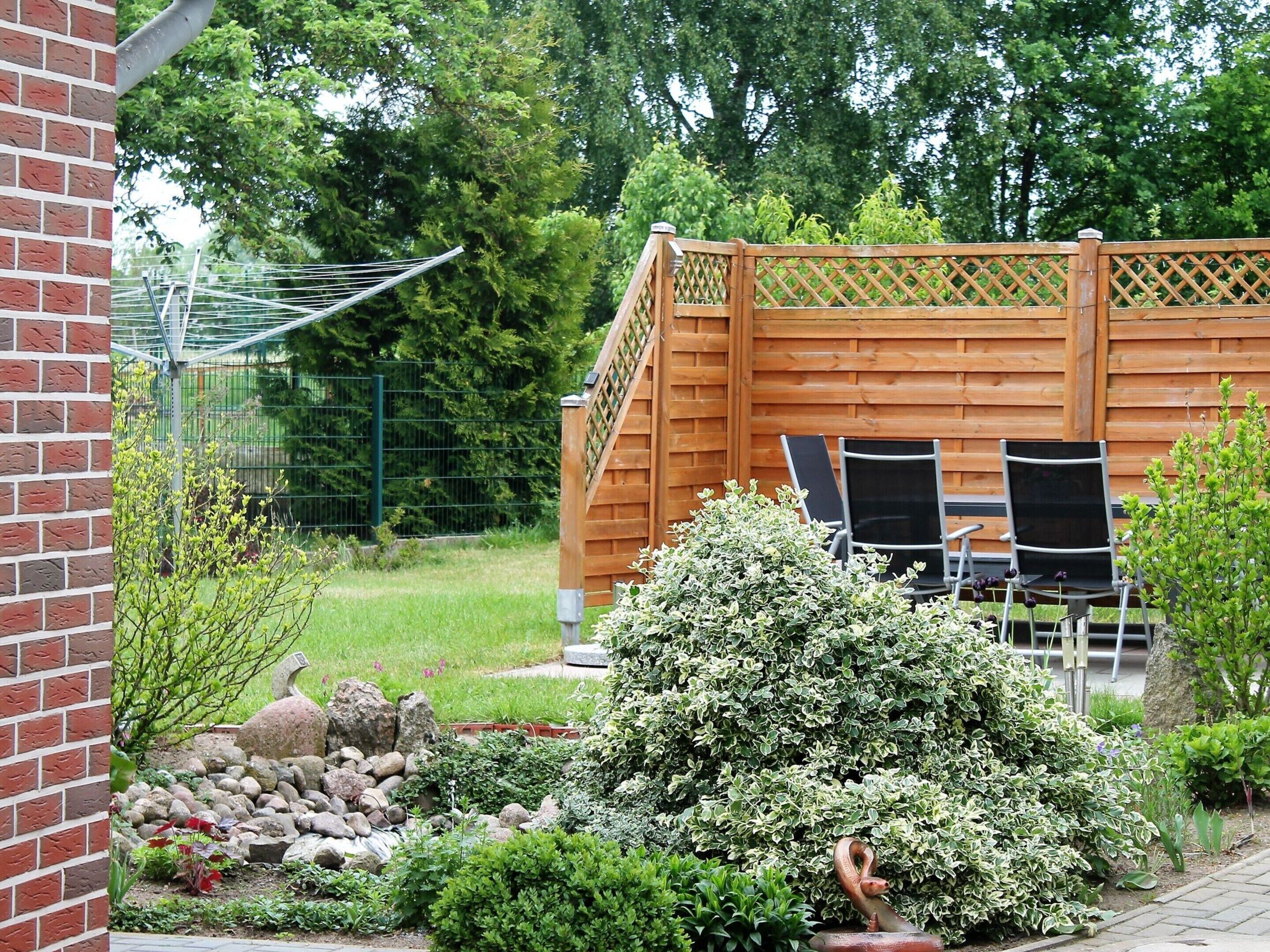 Una terraza de madera con dos sillas, rodeada por un jardín con diversas plantas y un muro de ladrillo.