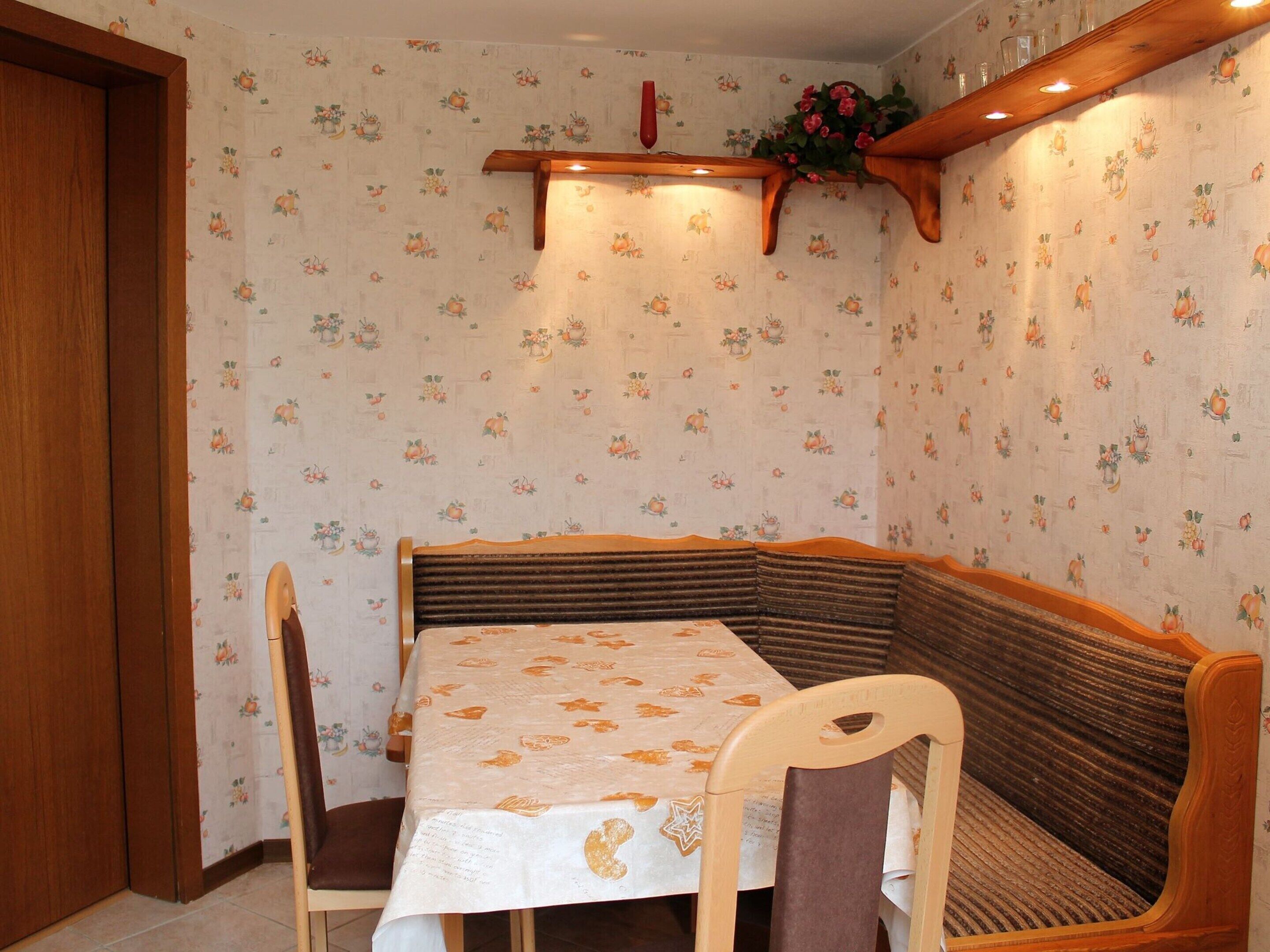 A dining area with a wooden bench, a table with a floral tablecloth, and a wall with a fruit pattern.