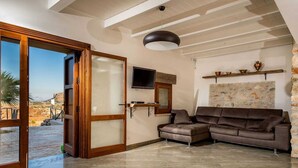 A spacious living area with a large sliding glass door leading to an outdoor patio, a brown leather sofa, a mounted flat-screen TV, and a stone wall with a decorative shelf. - Villa in Sicily Near Guidaloca Bay (Castellammare del Golfo)