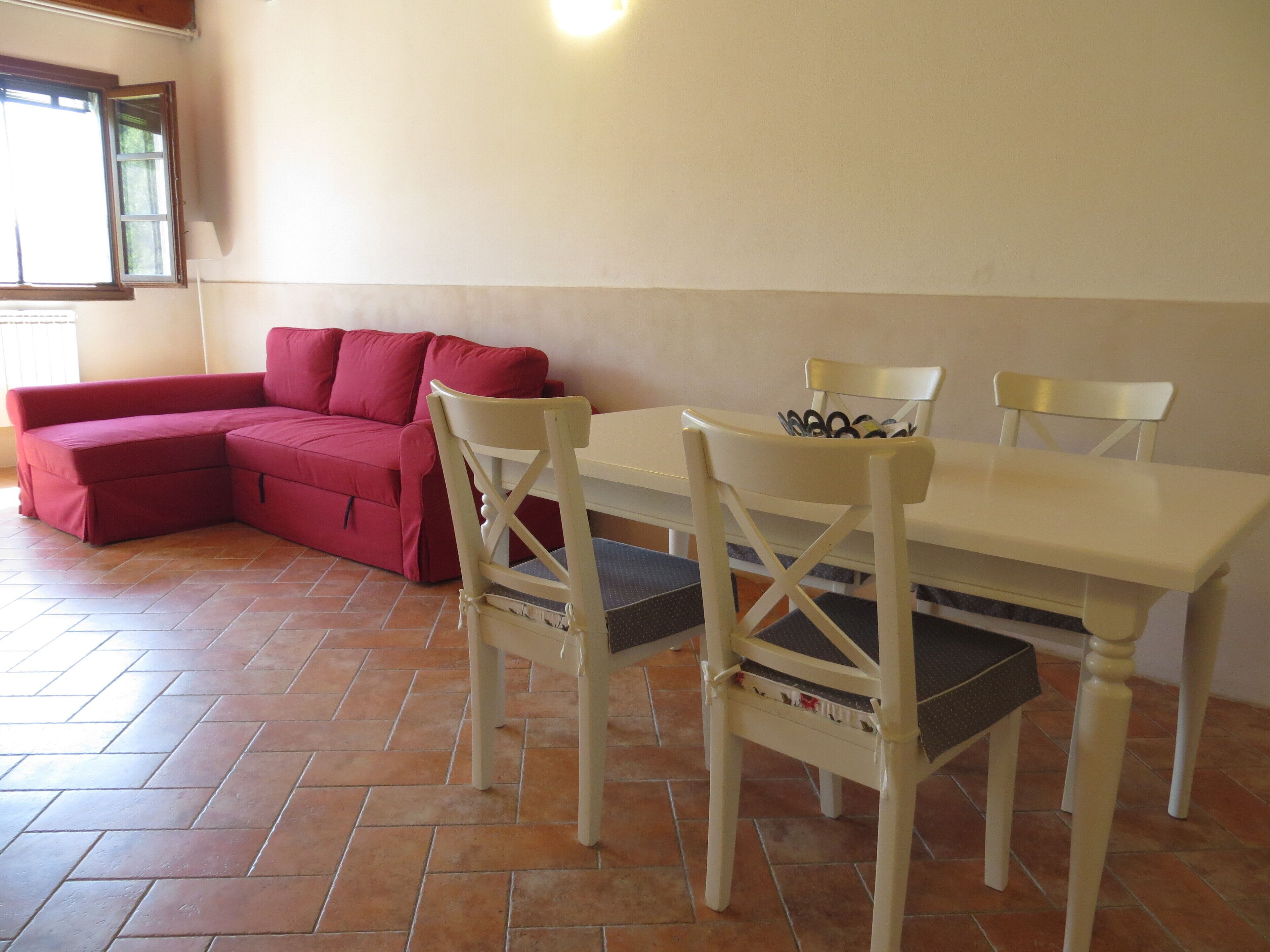 A room with a red sectional sofa, a white dining table with four chairs, and a window with a view of trees.