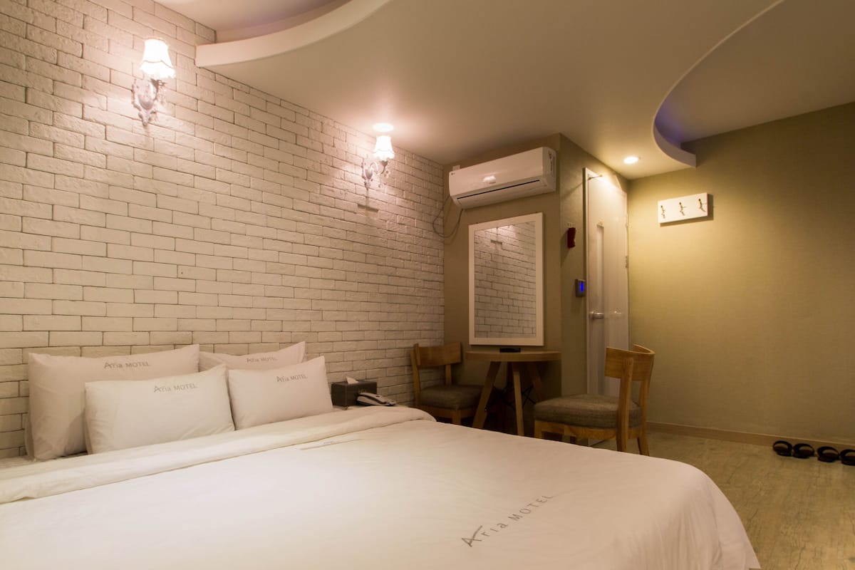 A hotel room with a brick wall, a bed with white bedding, a desk with a chair, and an air conditioning unit.