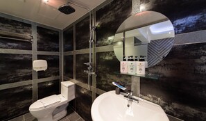A modern bathroom with a toilet, sink, and shower. There is a mirror with a glass shelf holding toiletries. The walls have a dark, textured finish. - Aria Motel (Busan)