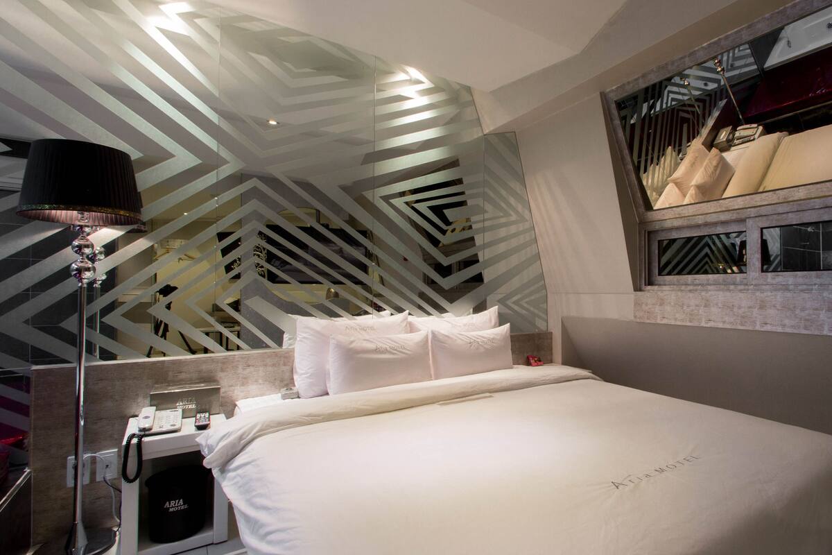 A modern hotel room with a bed, a nightstand with a lamp, a mirror, and a geometric patterned wall.