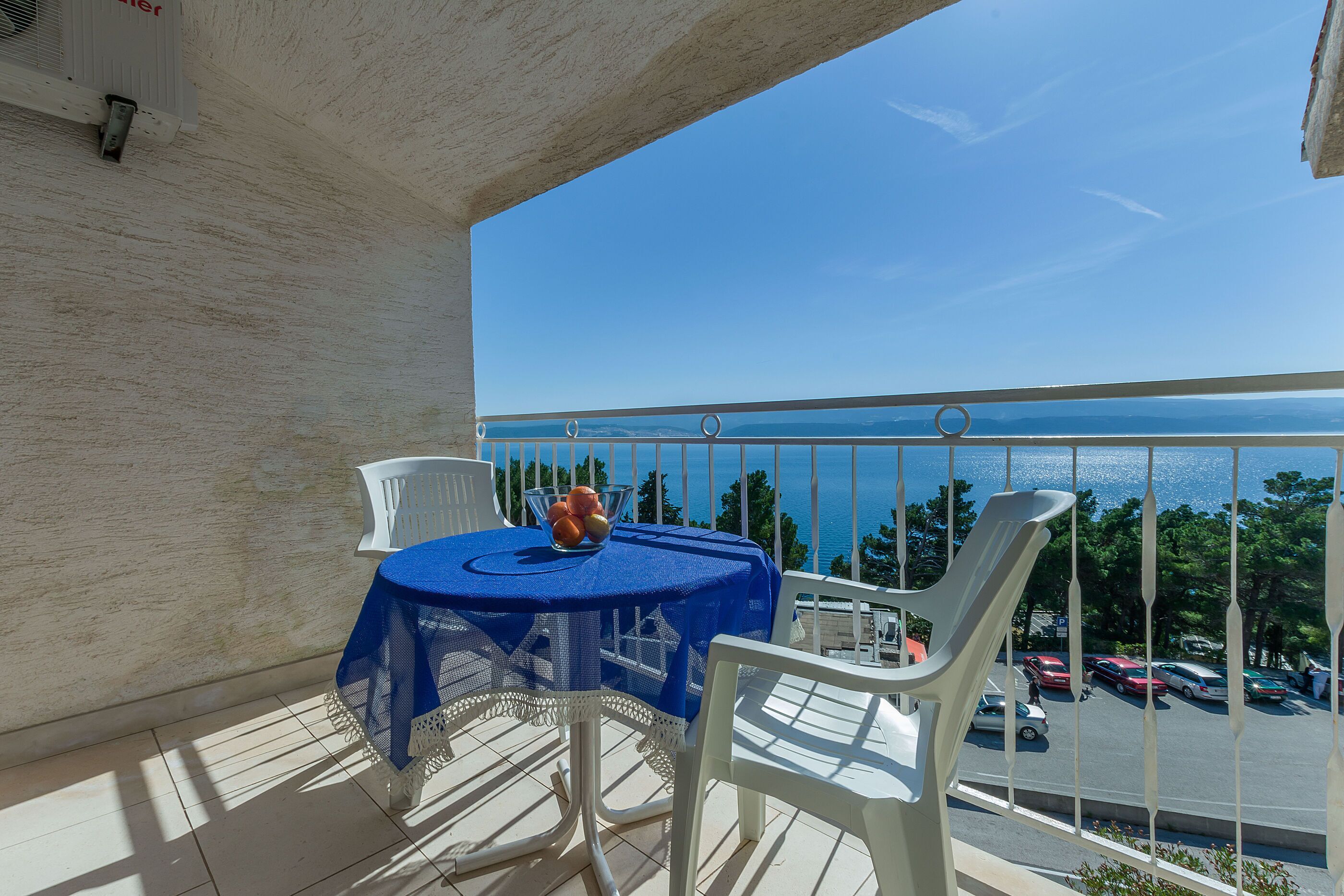 Un balcon avec une table et des chaises, donnant sur un parking et une étendue d’eau.