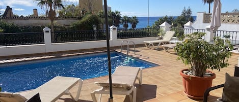 A poolside area with a swimming pool, lounge chairs, a potted plant, and a view of the ocean.