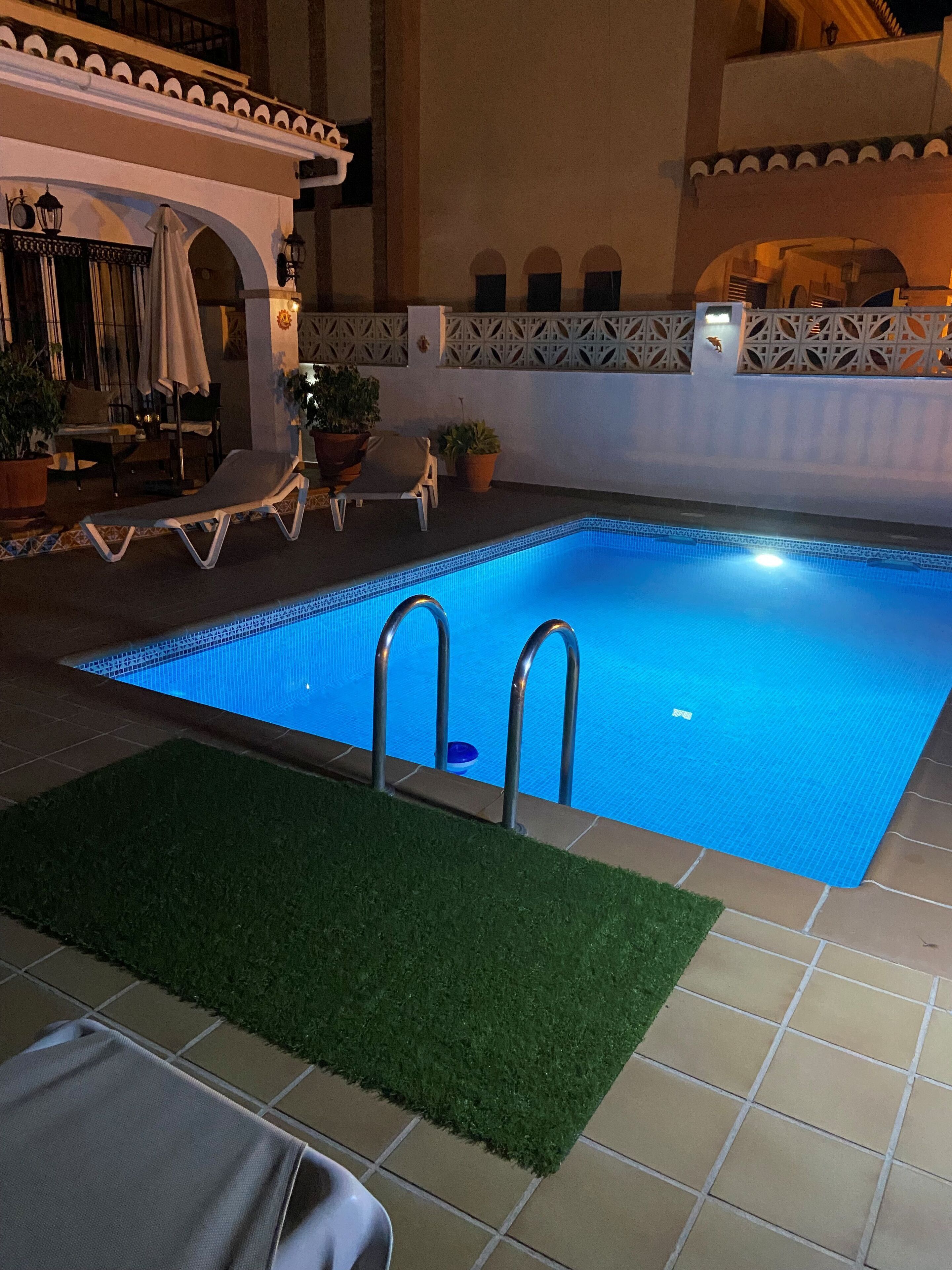 Indendørs swimmingpool med fliserede vægge, et loungeområde med stole og parasoller, samt plantekasser med planter.