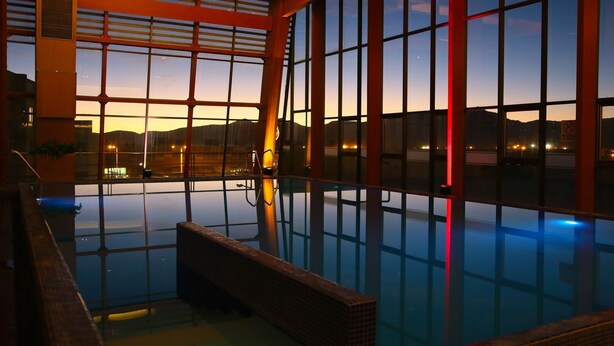 Una amplia piscina interior con paredes de vidrio transparente, un solárium de madera y vistas al atardecer y al paisaje urbano.