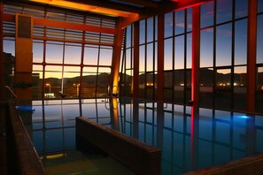 Une grande piscine intérieure aux parois en verre transparent, dotée d’une terrasse en bois et offrant une vue sur le coucher du soleil et le paysage urbain.