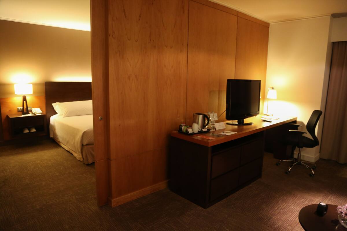 A hotel room with a bed, a desk with a computer, a chair, a nightstand with a lamp, and a wooden wardrobe.
