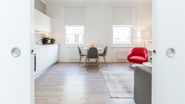 Une cuisine moderne avec des meubles blancs, une table à manger entourée de quatre chaises et un fauteuil rouge dans l’espace salon.