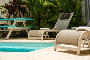 Uma área à beira da piscina com espreguiçadeiras e uma mesa, rodeada por plantas tropicais exuberantes.