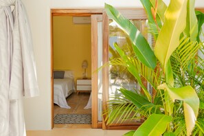 Quarto com cama, mesa de cabeceira, abajur e vista para uma varanda com plantas.