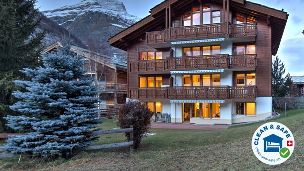 A multi-story wooden building with balconies, a snow-covered mountain in the background, and a logo for "Clean & Safe" in the bottom right corner.