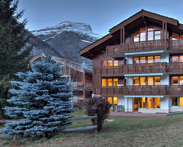 A multi-story wooden building with balconies, a snow-covered mountain in the background, and a logo for "Clean & Safe" in the bottom right corner. - Apartment for 2 to 4 people in Zermatt (Zermatt)