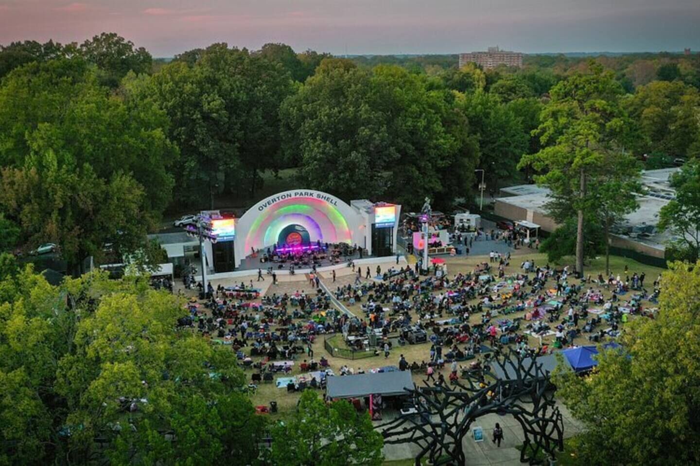 A large outdoor amphitheater with a stage and audience seating, surrounded by trees.